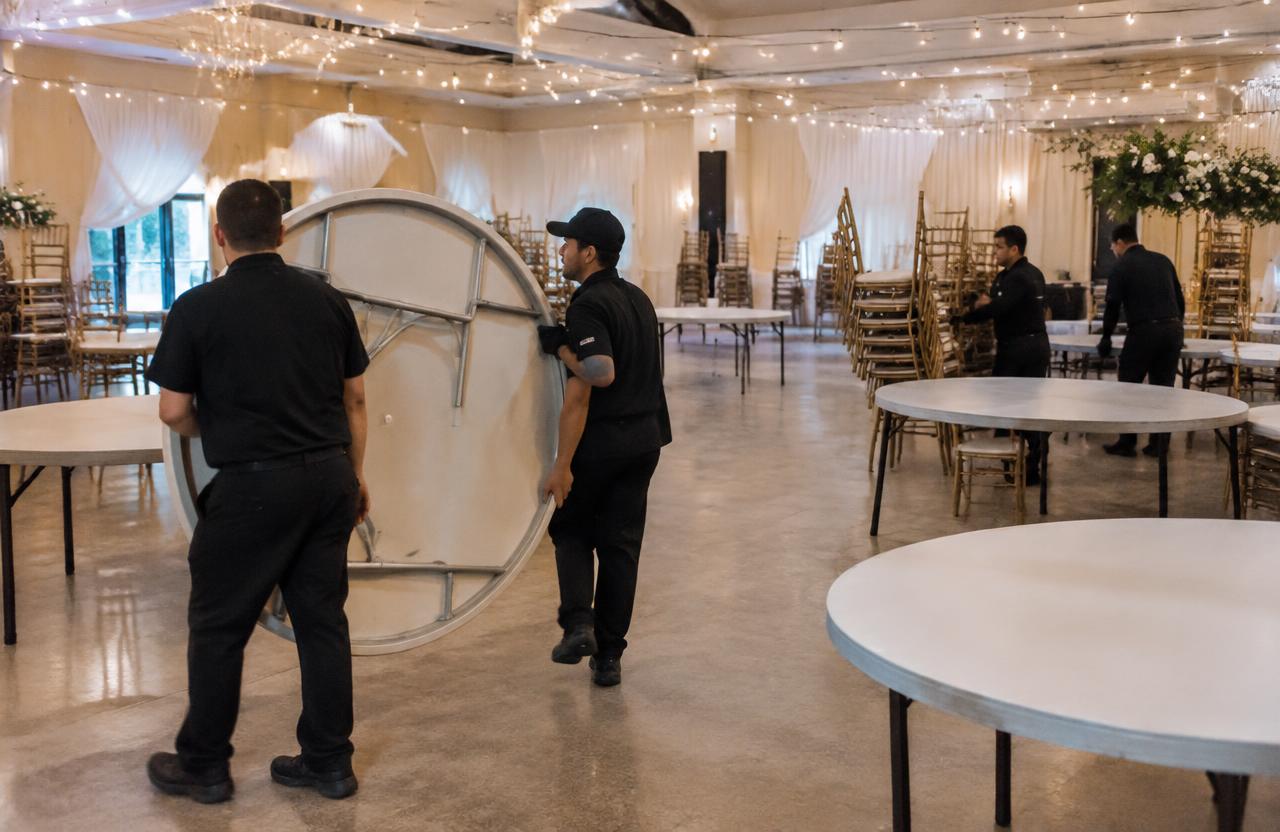 Crew carrying a folding round table through a venue with stacked chairs ready for breakdown
